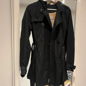 Svart trenchcoat med rutigt foder - Snygg svart trenchcoat med klassiskt rutigt foder. Jackan har långa ärmar och knappar framtill samt ett bälte i midjan för en elegant look. 