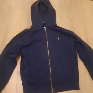 Säljer en snygg mörkblå hoodie från Ralph Lauren med dragkedja och den klassiska loggan på bröstet. Perfekt för kyliga dagar och har en skön passform. Den är i bra skick och redo för en ny ägare!