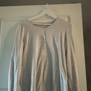 Beige kofta med knappar - Säljer en enkel och stilren grå/beige kofta med knappar framtill. Perfekt för lager-på-lager under höst och vår. Koftan har långa ärmar och är i bra skick. Passar både till vardags och lite finare tillfällen.