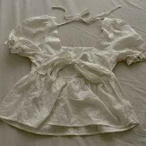 Vit blus med knytband - Söt vit blus med puffiga kortärmar och knytband i ryggen. Perfekt för sommardagar eller en avslappnad vårlook. Blusen har en luftig passform och volangdetaljer vid ärmsluten.