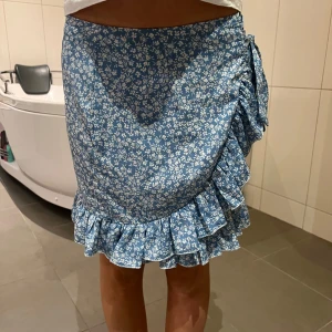 Blå blommig kjol med volang - Superfin blå blommig kjol med volangdetaljer och knytning i sidan. Perfekt för sommardagar eller en mysig vårutflykt. Kjolen har en omlott-design som ger en söt och feminin look. Passar perfekt till en enkel topp och sandaler! Säljes då den inte kommer till användning löngre 