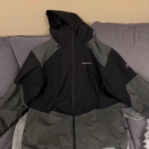 Svart och mörkgrå jacka från Peak Performance - Säljer en snygg svart och mörk grå  jacka från Peak Performance. Den har en praktisk dragkedja och huva, perfekt för höst,vinter och vår. Jackan är i bra skick och passar för både vardag och utomhusaktiviteter. Storleken är herr xxl. Perfekt för dig som vill ha en stilren och funktionell jacka!