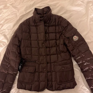 Brun quiltad jacka från Moncler - Säljer en snygg brun quiltad jacka från Moncler. Den har en klassisk design med långa ärmar och praktiska fickor. Perfekt för höst och vinter, håller dig varm och stilren. Jackan har en diskret logga på ärmen och stängs med knappar.