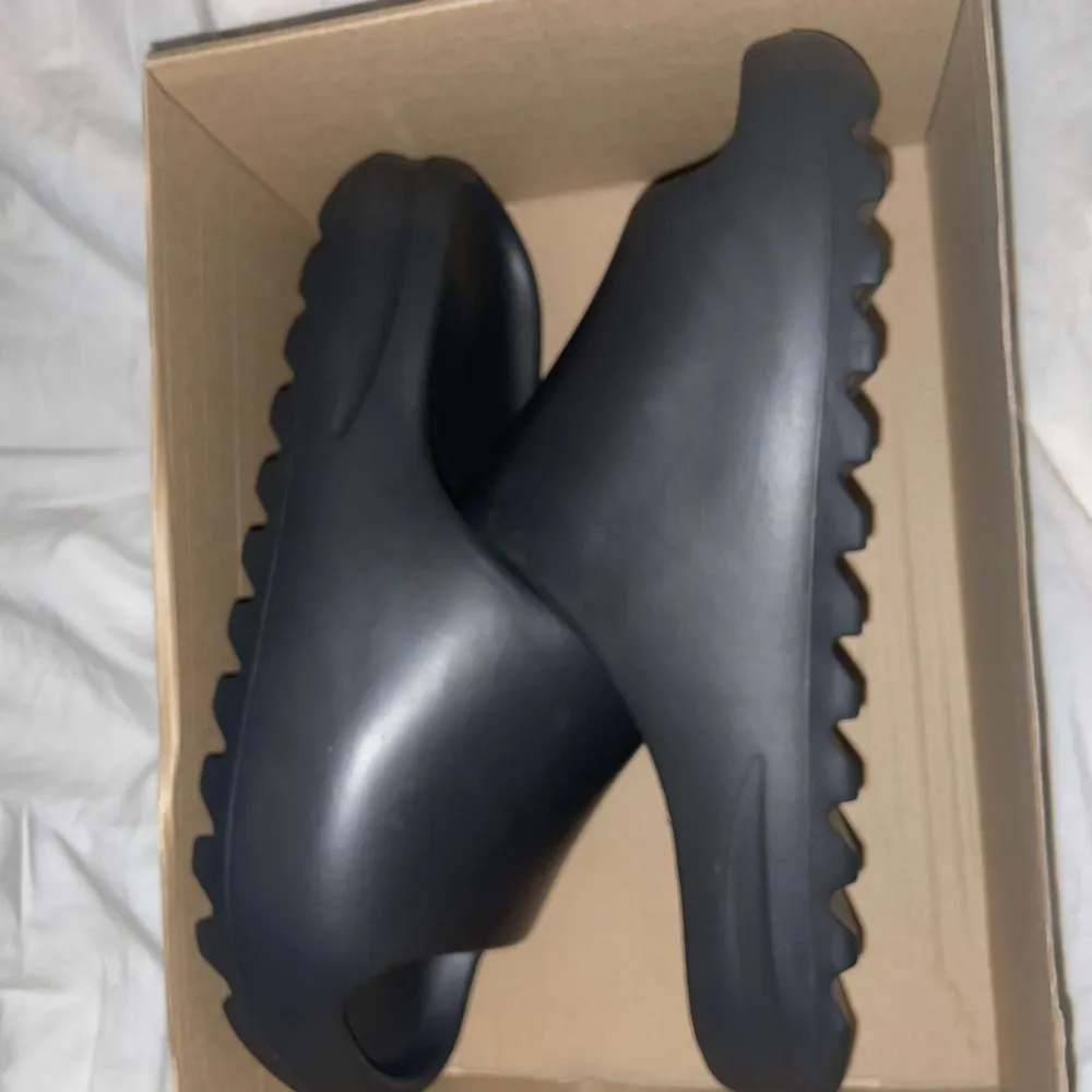 Säljer ett par svarta Yeezy Slide från Adidas i storlek herr 45. De är i syntetmaterial och har en modern, minimalistisk design med en bekväm passform. Perfekta för sommaren med sin öppna tå och räfflade sula för extra grepp.. Skor.