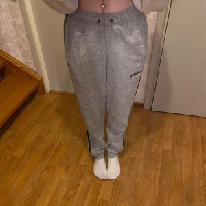 Grå mjukisbyxor från Adidas - Säljer ett par grå mjukisbyxor från Adidas i mycket bra skick. De har en normal passform med klassiska svarta ränder längs sidorna och en elastisk midja. Perfekta att ha som mjukisbyxor eller träning. Notera att smörerna saknas.