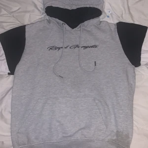 Grå hoodie med tryck - Säljer en grå hoodie med svart text på bröstet. Den har en skön luva och en stor ficka framtill. Ärmarna är i en cool kombination av svart och grått med en vit rand. Perfekt för en avslappnad stil!