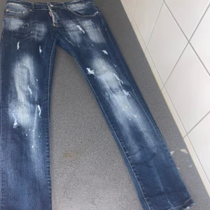 Blå jeans från Dsquared2 - Säljer ett par coola blå jeans från Dsquared2 i storlek 46 . De har en unik distressed look med slitningar och färgstänk som ger en edgy stil. Perfekta för en avslappnad men trendig outfit. Tillverkade i Italien med hög kvalitet.