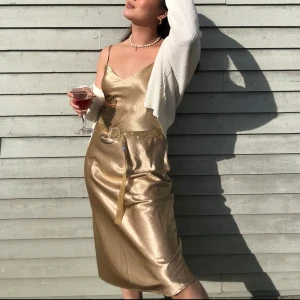 Guldig klänning - Säljer en elegant guldig klänning med smala axelband som går att justera och v-ringad hals. Perfekt för festliga tillfällen. Klänningen har en glansig finish, i bra skick men kan ha lite små skavanker som endast syns om man kollar väldigt nära (se bild 4)