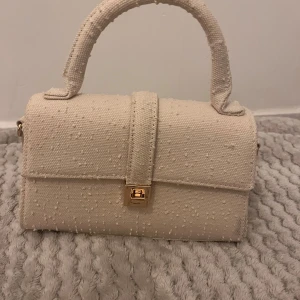 Beige handväska med axelrem - Säljer en superfin beige handväska i mycket bra skick. Den har en elegant struktur med små prickar och en snygg guldfärgad låsdetalj. Väskan kommer med en avtagbar axelrem, perfekt för både vardag och fest. Den är både stilren och praktisk!