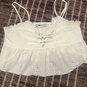 Vit somrig topp med spetsdetaljer - Säljer en superfin vit topp från New Yorker i storlek S, fast den passar xs också. Den har smala axelband och vacker spetsdetaljering. Toppen är croppad och har en söt knytning framtill. 