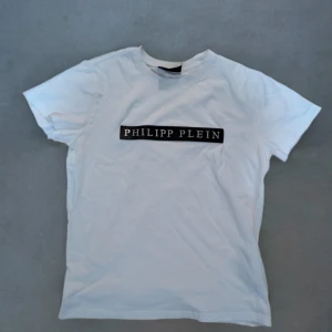 Vit t-shirt från Philipp Plein - Säljer en stilren vit t-shirt från Philipp Plein i mycket bra skick. Den har ett svart tryck med märkesnamnet på bröstet. Perfekt för en casual look eller att klä upp med en snygg jacka. Passar till alla säsonger och är ett måste i garderoben!