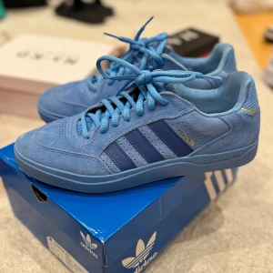 Blå Adidas sneakers - Säljer ett par snygga blå Adidas sneakers i mocka. De har de klassiska tre ränderna i en mörkare blå nyans och snörning framtill. Perfekta för både vardag och träning. Skorna är i mycket bra skick och kommer i originalkartong.