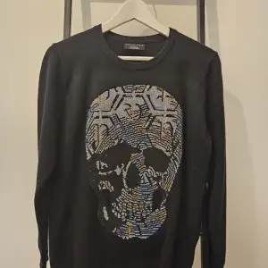 Säljer en svart tröja från Philipp Plein med en cool dödskalle-design i olika färger på framsidan. Tröjan har långa ärmar och är perfekt för en edgy look. Passar både till vardags och fest!