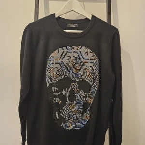 Svart tröja med dödskalle från Philipp Plein - Säljer en svart tröja från Philipp Plein med en cool dödskalle-design i olika färger på framsidan. Tröjan har långa ärmar och är perfekt för en edgy look. Passar både till vardags och fest!