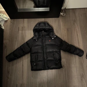 Svart dunjacka från Polo Ralph Lauren Dam - Säljer en snygg svart dunjacka från Polo Ralph Lauren. Jackan har en klassisk pufferstil med huva och dragkedja framtill. Den är dekorerad med det ikoniska röda Polo-logotypen på bröstet. Perfekt för kalla vinterdagar! 🖤jackan enda nackdel är att den har smink fläckar men det går bort med en enkel kemtvätt. Kostar ca 250-400kr så blir den som ny. Nypris 6,700kr 