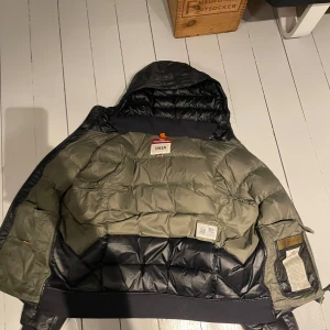 Parajumper jacka - Säljer en snygg och varm svart dunjacka från Parajumpers, modell Sheen. Jackan har en quiltad design med en cool patch på ärmen. Perfekt för kalla vinterdagar. Den är i mycket bra skick och har en skön passform. Huvudfärgen är svart med inslag av grönt på insidan.