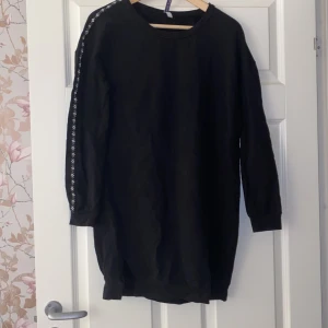 Svart klänning från Miss E Collection - Säljer en svart klänning från Miss E Collection. Den har långa ärmar med ett snyggt mönster längs sidorna. Perfekt för en avslappnad stil eller att mysa i hemma. Passar bra till leggings. Köpt i Nederlängderna.