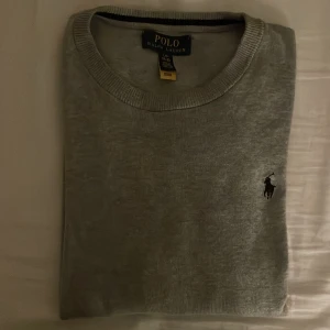 Grå tröja från Polo Ralph Lauren - Säljer en stilren grå tröja från Polo Ralph Lauren i storlek L i barn. Tröjan är i mycket bra skick och har en klassisk design med rund hals och det ikoniska logotypbroderiet på bröstet. Perfekt för både vardag och lite finare tillfällen. Passar bra till  alla årstider.