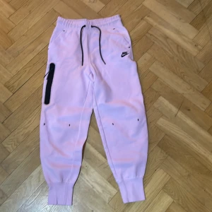 Rosa mjukisbyxor från Nike - Säljer ett par superbekväma rosa mjukisbyxor från Nike. De har en elastisk midja med snörning och en praktisk ficka på sidan. Perfekta för en avslappnad dag hemma eller en snabb tur till affären. 🩷