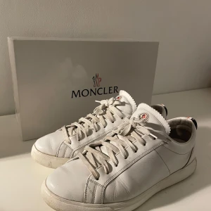 Vita sneakers från Moncler - Säljer ett par stilrena vita sneakers från Moncler i skinn. De har klassisk snörning och en bekväm passform. Perfekta för både vardag och festliga tillfällen. Skorna kommer med originalkartong och dustbag. Använda men i bra skick. Storlek 41 men passar 42 och 42,5