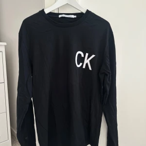 Svart långärmad tröja från Calvin Klein - Säljer en stilren svart långärmad tröja från Calvin Klein med vita initialer 'CK' på bröstet. Perfekt för en casual look och passar till det mesta. Tröjan är i bra skick och gjord av mjukt material för extra komfort.
