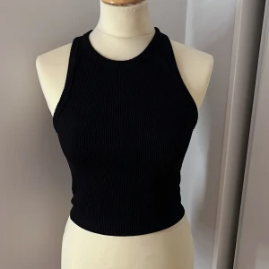 Svart ribbad linnetopp - Snygg svart ribbad linnetopp i ett stretchigt material. Perfekt för sommardagar eller som ett lager under en jacka. Den är ärmlös och har en tight passform som framhäver figuren. Ett måste i garderoben för en stilren look!