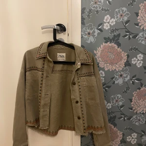 Grön jacka från Zara - Säljer en superfin beige jacka från Zara i storlek XS. Jackan har snygga broderade detaljer och knappar framtill. Perfekt för vår och höst med sin croppade stil och långa ärmar. Passar både till vardags och fest!