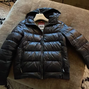 Svart pufferjacka från Parajumpers - Säljer en snygg svart pufferjacka från Parajumpers i modellen Sheen. Jackan har en quiltad design med dragkedja och en cool patch på ärmen. Perfekt för höst och vinter, håller dig varm och stilren. Passar både till vardags och lite finare tillfällen. 🖤några defekter som jag har sytt som man ser knappt.