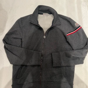 Grå Moncler cardigan  - Säljer en snygg grå tröja från Moncler i storlek M. Den har en dragkedja framtill och ett coolt Moncler-märke på ärmen med röd och vit rand. Perfekt för både vardag och lite finare tillfällen. Superbekväm och stilren!