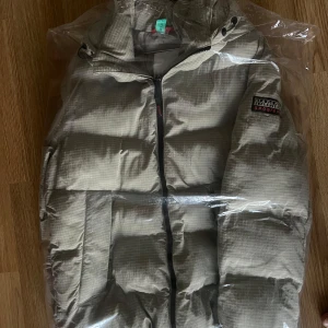 Beige dunjacka från Napapijri - Säljer en snygg beige dunjacka från Napapijri i storlek S. Jackan har ett diskret rutmönster och är perfekt för kalla vinterdagar. Den har en dragkedja framtill och en mysig huva. Perfekt för både vardag och utomhusaktiviteter! Nypris 5500kr använt en vinter. 