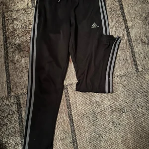 Svarta träningsbyxor från Adidas - Säljer ett par svarta träningsbyxor från Adidas i storlek 152. Byxorna har de klassiska tre ränderna längs sidorna och är tillverkade i ett lätt och bekvämt material. Perfekta för träning eller en avslappnad dag. De har en elastisk midja med snörning för bästa passform och fickor med dragkedja. 