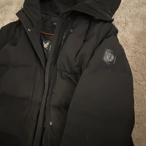 Svart dunjacka från Hollies - Säljer en svart dunjacka från Holies i använd skick,fodret insidan av armhålan har gått sönder kan sys ihop. Jackan har en praktisk huva och stängs med både dragkedja och knappar. Perfekt för kalla vinterdagar! Den har långa ärmar och två fickor med dragkedja framtill. En stilren och varm jacka för vintern.