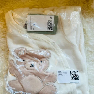 Vit babytröja med nallemotiv från H&M. Oanvänd.  - Supergullig vit babytröja från H&M med ett stort nallemotiv på framsidan. Perfekt för små barn i åldern 2-3 år. Tröjan är i nytt skick och kommer i originalförpackning. Den är mjuk och bekväm, idealisk för både lek och mysiga stunder.