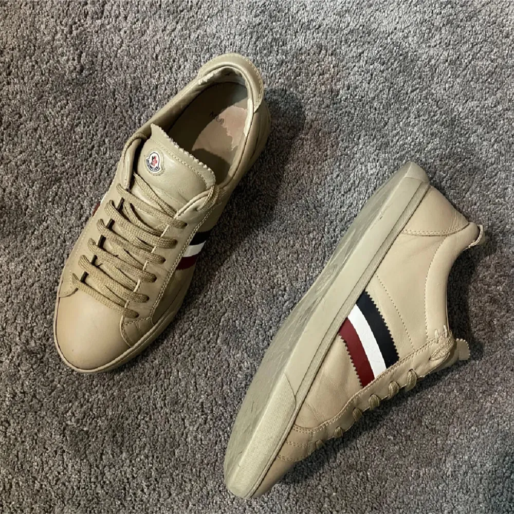 Snygga beige sneakers från Moncler i skinn med klassiska ränder i rött, vitt och svart på sidan. De har en stilren design med snörning och Moncler-logga på plösen. Perfekta för både vardag och festliga tillfällen. Skorna är i mycket bra skick och redo att bäras med stil! Storlek 41,5 men passar en storlek större!. Skor.