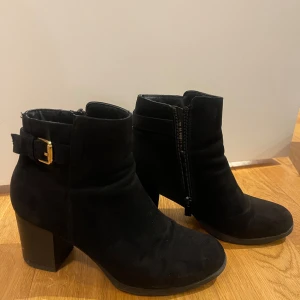 Svarta boots med klack  - Snygga svarta boots i mocka med klack och ett dekorativt spänne på sidan. De har en praktisk dragkedja för enkel på- och avtagning. Perfekta för höst och vinter.