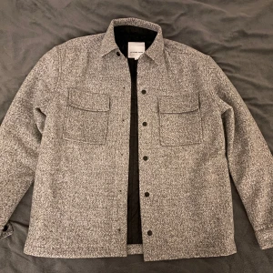 Lindbergh Overshirt - Tja! Säljer min Lindbergh Overshirt i L då jag fick den som present och aldrig använt den. Beskrivning av jackan: Overshirt from Lindbergh White in a marled grey woollen fabric. The shirt is made in a simple design with two hidden side pockets and two breast pockets with press studs. Made in relaxed fit, which is a bit tighter than regular fit but still provides room for movement.