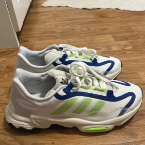Vita och blå sneakers från Adidas - Snygga vita sneakers från Adidas med blå och limegröna detaljer. Skorna har en modern design med snörning och en bekväm sula. Perfekta för en sportig look.