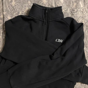 Svart tröja med dragkedja från ICANIWILL - Säljer en stilren svart tröja från ICANIWILL med halv dragkedja och hög krage. Tröjan har långa ärmar och en liten logga på bröstet. Passar bra till både träning och vardag.
