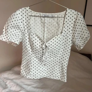 Vit prickig blus från NA-KD - Charmig vit blus med svarta prickar från NA-KD. Blusen har korta puffärmar och en söt knytning framtill. Perfekt för en somrig look med sin lätta och luftiga design. Toppen har även ett litet hål under armhålan men det är inget man märker av☀️