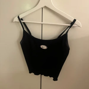 Svart linnetopp från Nike - Snygg svart linnetopp från Nike med smala axelband och en liten logga framtill. Perfekt för en sportig look eller som en del av en avslappnad outfit. Den ska såklart tvättas innan eftersom den har del fläckar🥰