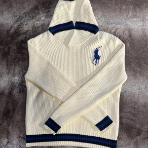 Ralph Lauren - Snygg och välbehållen Ralph Lauren stickad tröja i ljusbeige/vit färg. Tröjan har en mocka-krage och blå/svarta ränder vid ärmsluten och nederkanten.  Tröjan är i fint skick men har en liten fläck på ena ärmen (se bild).