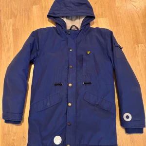Blå parkajacka från Lyle & Scott - Säljer en blå parkajacka från Lyle & Scott med huva och knappar framtill. Jackan har en gul logotyp på bröstet och flera praktiska fickor. Perfekt för kyligare dagar.