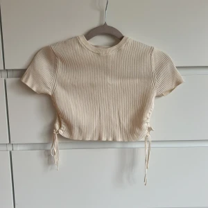 Beige ribbad topp från Zara - Säljer en beige ribbad topp från Zara med korta ärmar och knytning i sidorna. Toppen är croppad och har en tight passform.🤍använd endast 2 ggr!