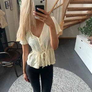 Gul blus med knytband - Säljer en söt gul blus med knytband i midjan och knappar framtill. Blusen har korta ärmar och v-ringad . Perfekt till våren/sommaren! Knappt använd!!