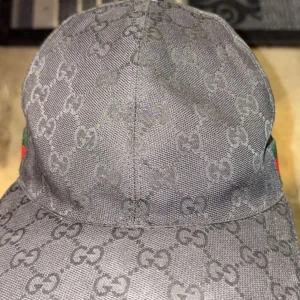 Svart gucci keps - Snygg svart keps från Gucci med det ikoniska monogrammönstret. Kepsen har en justerbar rem baktill i svart skinn och en grön och röd randig detalj på sidan. Perfekt för att ge din outfit en lyxig touch. Kvitto samt box kommer med🧢