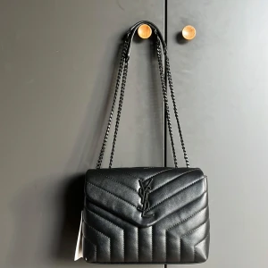 Svart axelväska från Saint Laurent - Snygg svart axelväska från Saint Laurent med quiltad design och kedjeaxelrem. Väskan har en elegant YSL-logga på framsidan. Perfekt för att ge din outfit en lyxig touch. Pris kan diskuteras, den är inte äkta!