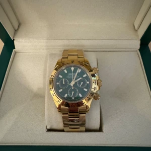 Seiko Daytona guld - Elegant guldfärgad klocka med svart urtavla och tre små urtavlor. Klockan har en robust länkarmband i guld och kommer i en lyxig grön förvaringsbox. Perfekt för den som vill ha en stilren och tidlös accessoar.