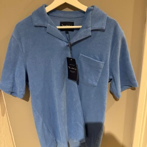 Bljusblå frotté piké från Bondelid - Säljer en stilren blå pikétröja från Bondelid. Tröjan har korta ärmar och en klassisk krage. Perfekt för en avslappnad och snygg look. Den har en bröstficka som ger en extra touch till designen.