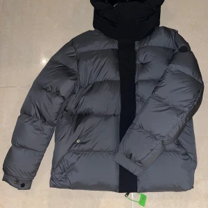 Grå dunjacka från Moncler - Säljer en snygg grå dunjacka från Moncler med hög krage och huva. Jackan har en stilren design med knappar och en diskret logga på ärmen. Perfekt för kyliga dagar. Jackan är helt ny och aldrig använd.