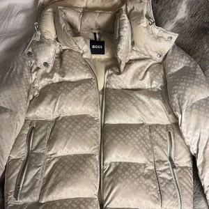 Beige pufferjacka från Boss - Snygg beige pufferjacka från Boss med diskret mönster och dragkedja framtill. Jackan har en hög krage och avtagbar huva